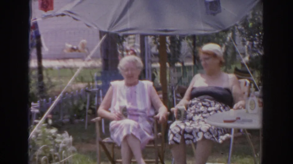 A 1966 Norfolk home-movie frame shows two seated older women under an umbrella in a sunny backyard garden.