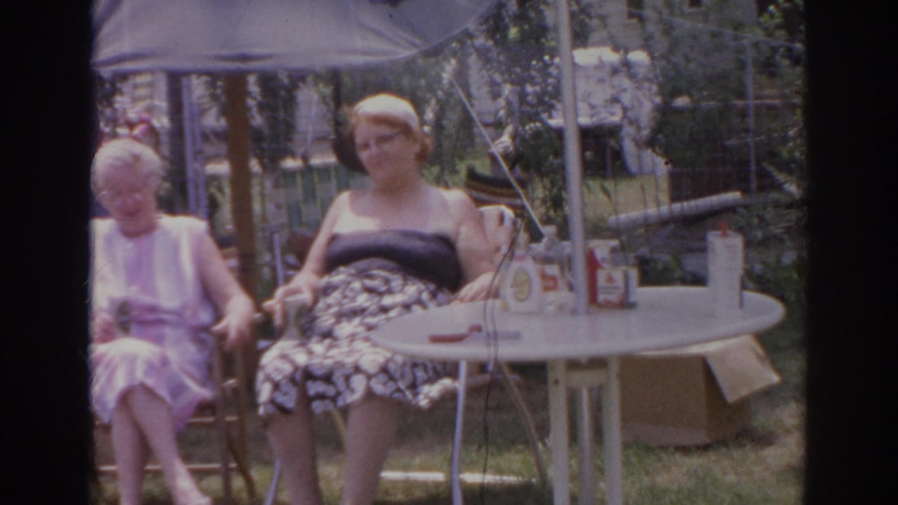 Two women sit outdoors in a 1966 Norfolk backyard, with bottles or condiments on a white patio table under an umbrella.