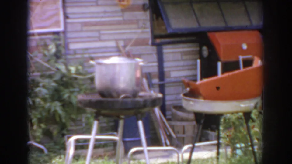 1966 Norfolk patio frame showing a metal pot on a grill, leafy plants, brick wall, and orange-and-white outdoor seating.