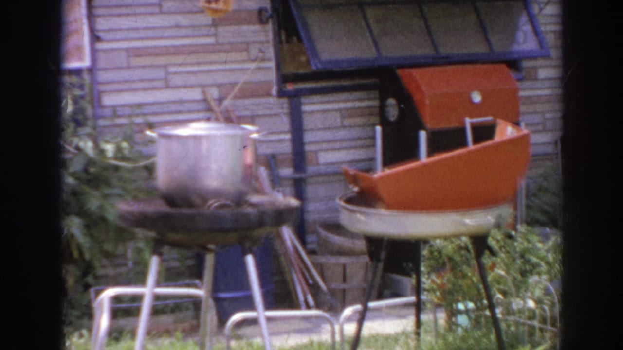 Backyard garden with orange kettle grill, metal pot, patio chairs, siding, and plants.