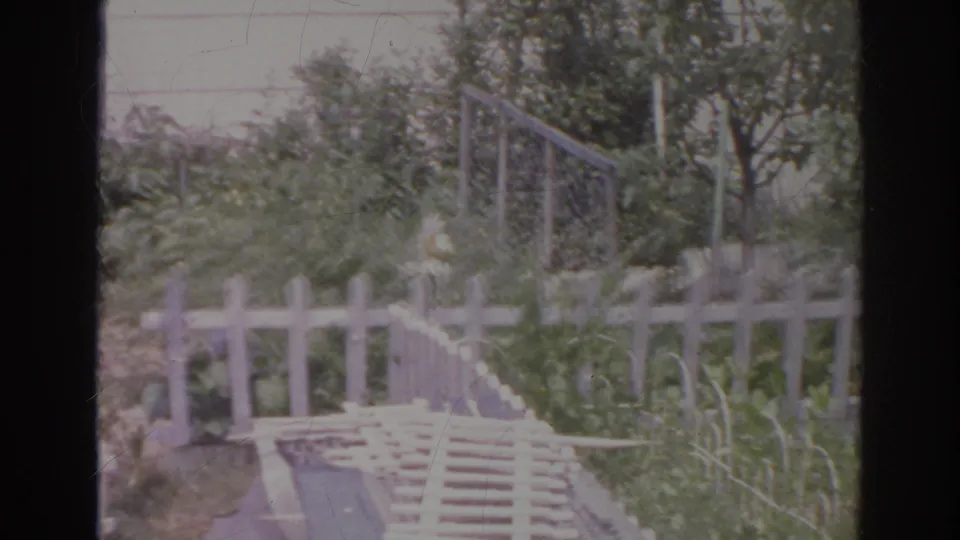 1966 Norfolk home-movie frame showing a cluttered garden corner with white fencing, railing, shrubs, and a small tree.