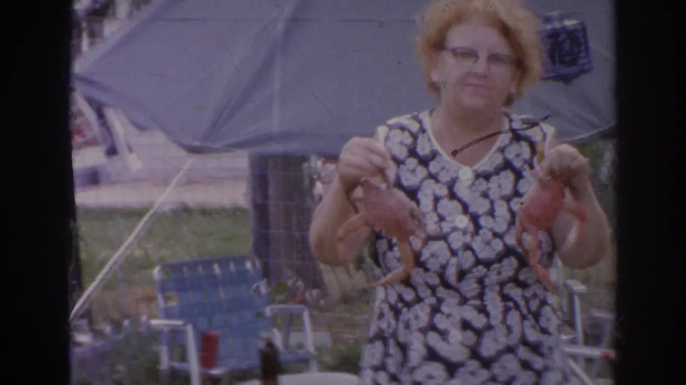 1966 Norfolk home-movie frame showing a woman in glasses and a patterned dress holding two red crabs in a backyard gathering area.