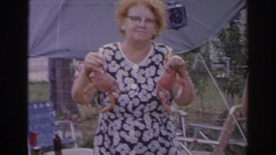 1966 Norfolk home-movie frame of a smiling woman holding two red crabs, with lawn chairs and a shade umbrella behind her.