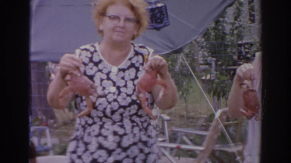 In 1966 Norfolk, Virginia footage, a woman in a black and white floral dress holds fresh crabs toward the camera in a backyard.