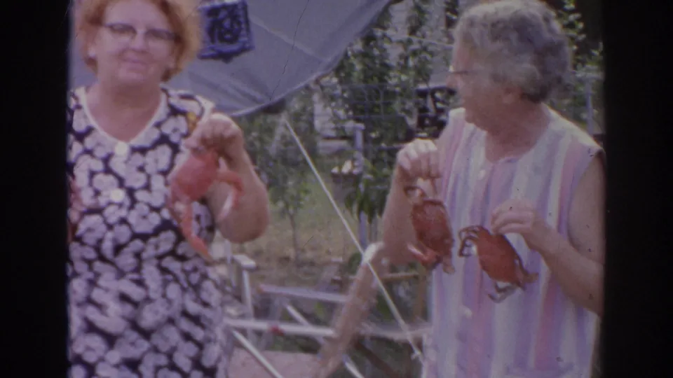 Two adult women in a Norfolk backyard hold crabs while standing near folding lawn chairs and a shade umbrella.