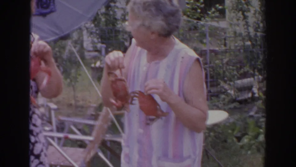 A gray-haired woman in a pink and white striped sleeveless top holds a crab beside another woman in the backyard scene.