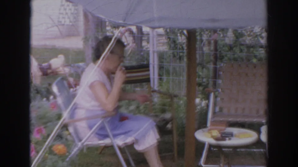 The woman remains seated under the umbrella, with garden flowers and a small round table visible beside her.