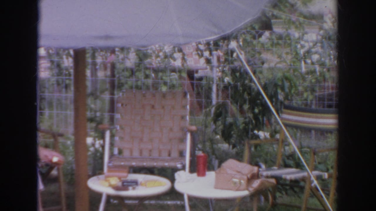 1966 Norfolk backyard patio scene with umbrella-shaded wicker seating, small tables, red thermos, brown purse, garden vines, and lawn.