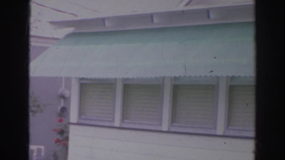 1966 Norfolk backyard view tilting toward a house exterior with mint-green awning, roofline, shuttered windows, and garden edge.