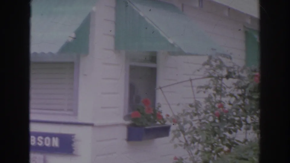 1966 Norfolk 8mm frame with green awnings, a partial DOBSON sign, red flowers, and leafy shrubs beside the facade.
