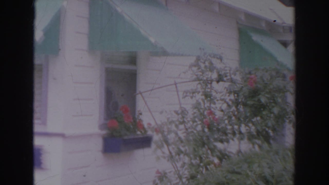 1966 Norfolk home-movie frame showing a white clapboard house with green awnings, red flowers, and foreground shrubs.