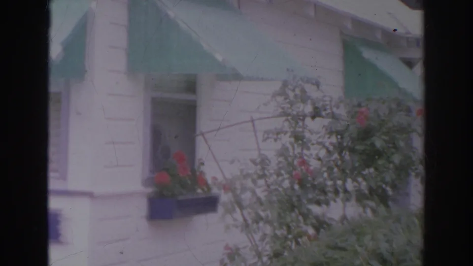 1966 Norfolk home-movie frame showing a white clapboard house with green awnings, red flowers, and foreground shrubs.