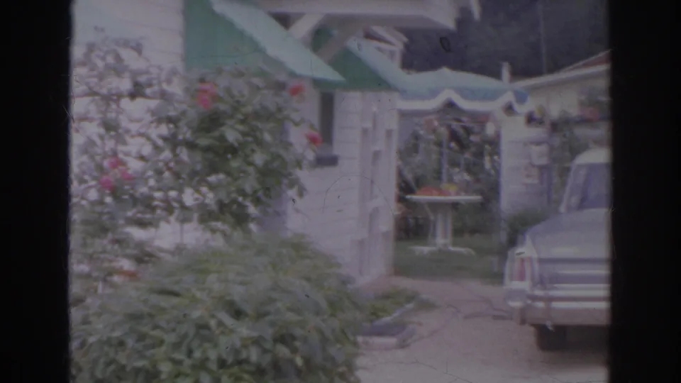 1966 Norfolk home-movie view across shrubs and a trellis toward a driveway with a partially visible classic sedan.