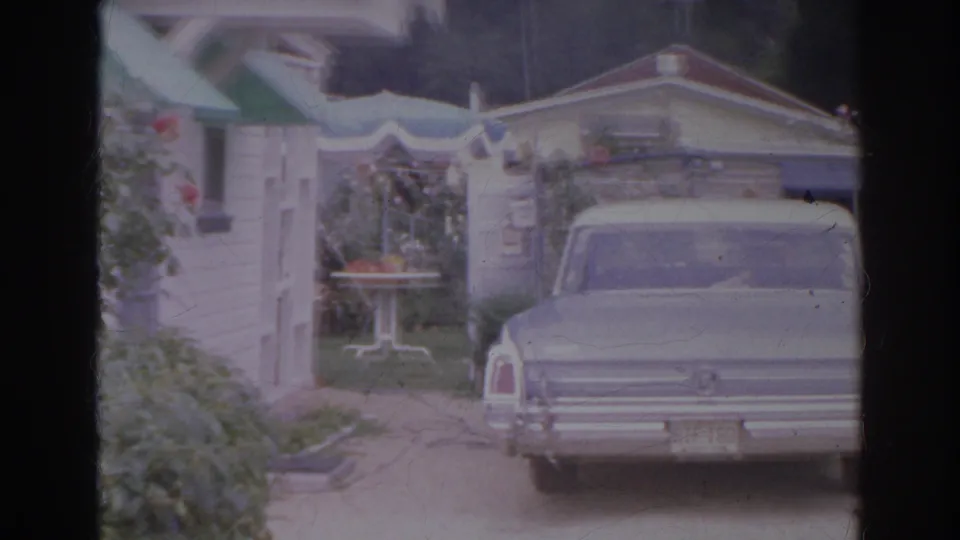 1966 Norfolk frame showing a classic 1960s sedan parked by a white house, garden trellis, shrubs, and patio furniture.