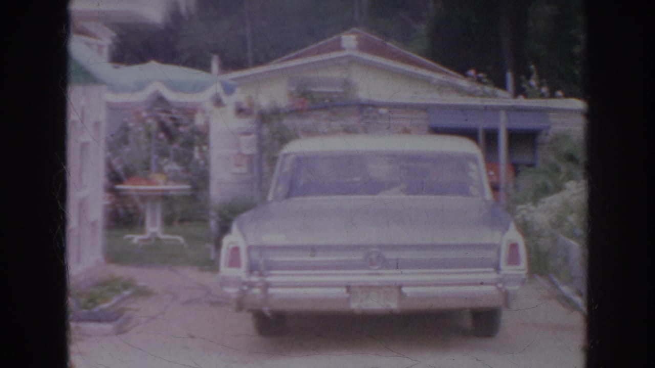 1966 Norfolk home-movie frame showing a pale sedan from the rear in a residential driveway near a house and garden area.