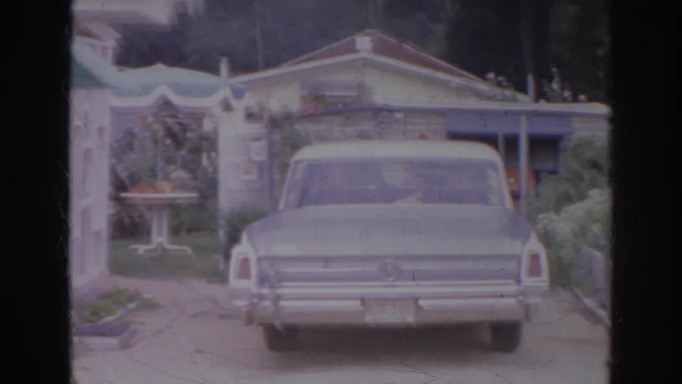 1966 Norfolk home-movie frame showing a pale sedan from the rear in a residential driveway near a house and garden area.