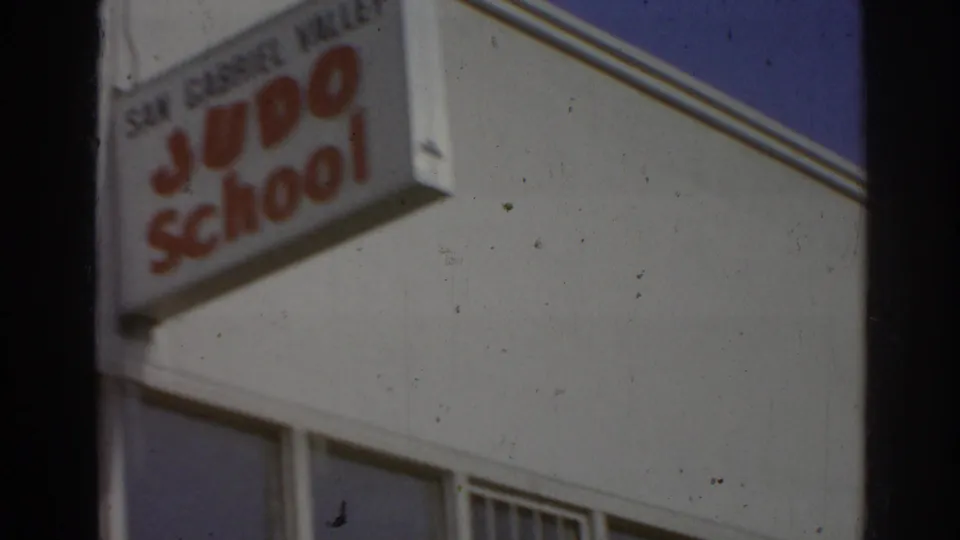 Soft 8mm frame showing the San Gabriel Valley Judo School sign on the exterior of a white building in California.