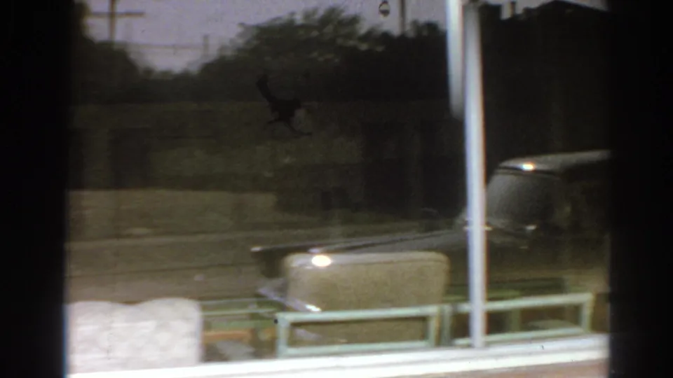1966 San Gabriel home-movie frame looking through a window toward lawn, a white pole, roadside greenery, and a passing car.