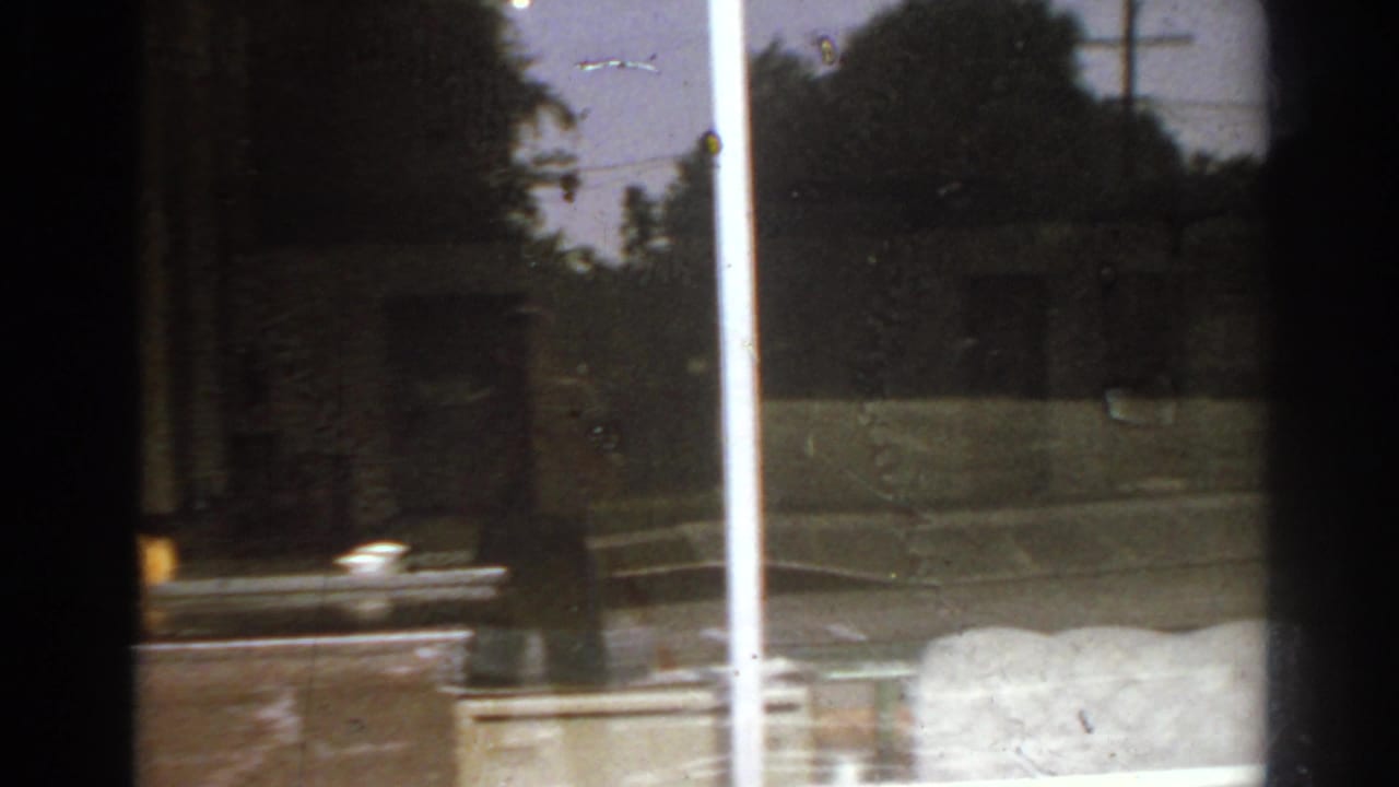 San Gabriel, California 1966 frame showing a window-side view of a white pole, exterior wall, greenery, and a car moving past.