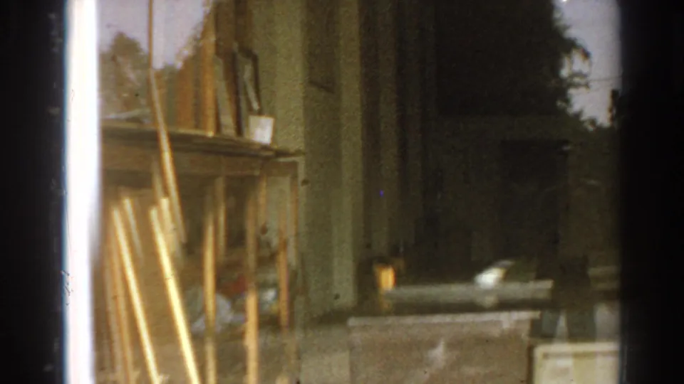 1966 home-movie frame from inside a San Gabriel house, showing wood interior detail, window edge, shrubs, and a car outside.