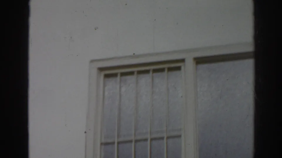 1966 San Gabriel home-movie frame showing a white building facade with a barred window and visible 8mm gate edges.