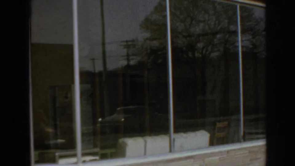 San Gabriel storefront windows reflecting trees, utility poles, and cars along the street in 1966 home-movie footage.