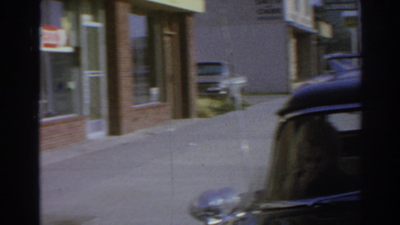 1966 San Gabriel home-movie frame showing a black car parked by a curb near a brick storefront and sunlit sidewalk.