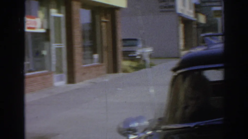1966 San Gabriel home-movie frame showing a black car parked by a curb near a brick storefront and sunlit sidewalk.