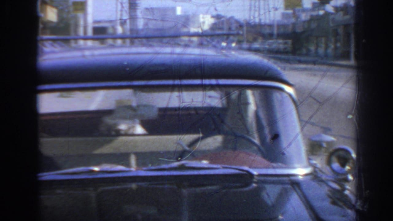 1966 San Gabriel footage shows the parked black car through glass, with a seated person faintly visible inside.