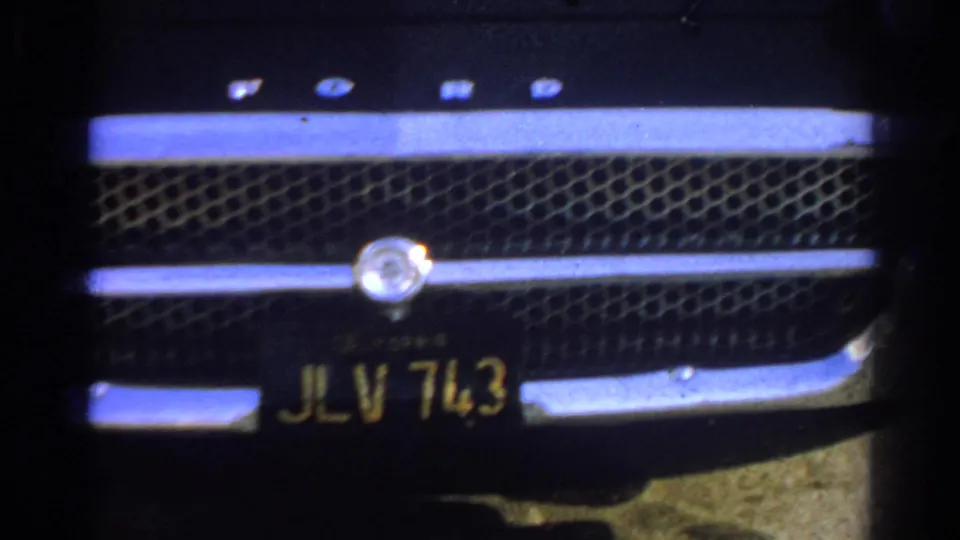 Close 1966 home-movie view of the black car's front grille, chrome bars, emblem area, and California-style black license plate.