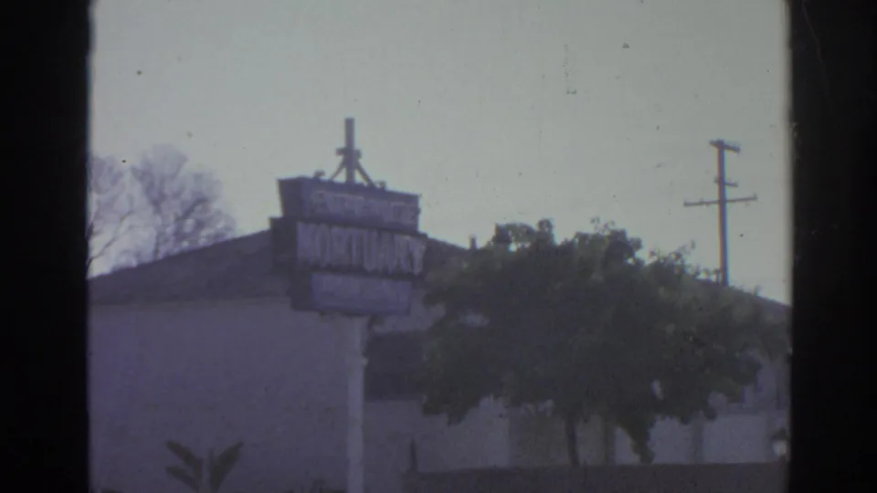 1966 San Gabriel street view showing a mortuary sign, low building roofline, trees, and utility poles under bright daylight.
