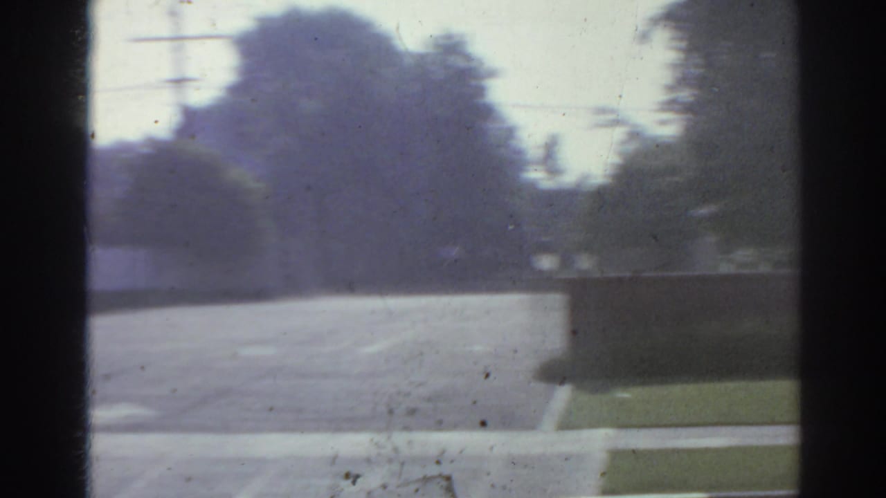 1966 San Gabriel home-movie frame showing a quiet road, sidewalks, trees, and low commercial buildings from a moving car.