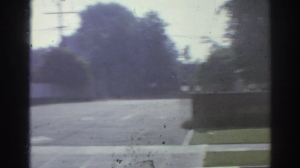 1966 San Gabriel home-movie frame showing a quiet road, sidewalks, trees, and low commercial buildings from a moving car.