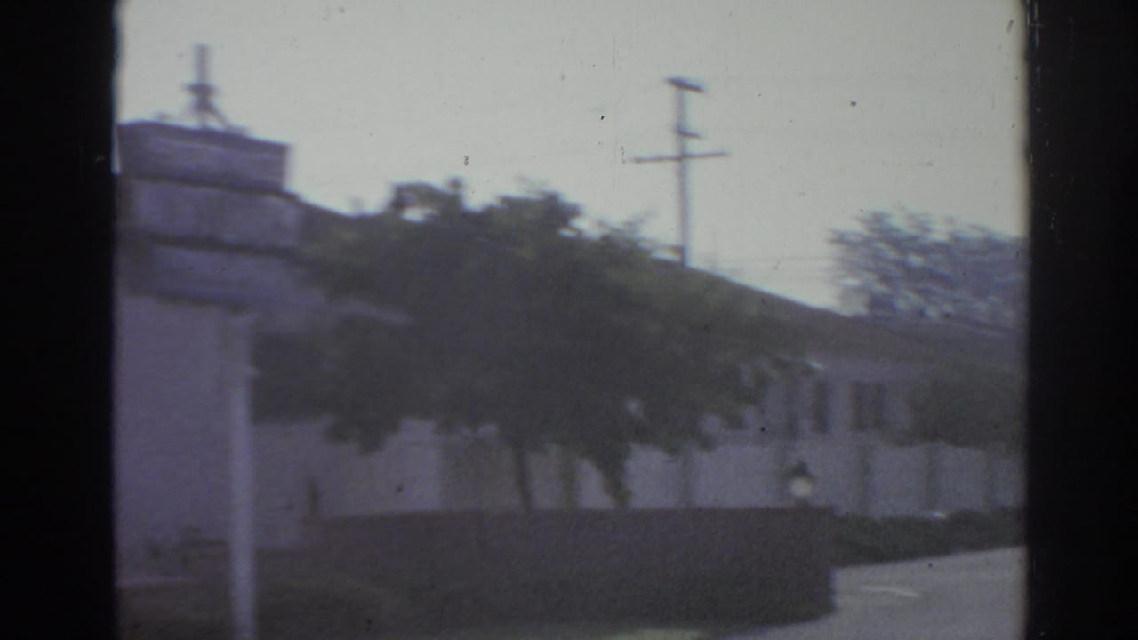 1966 San Gabriel street frame with a low commercial building, trees, utility pole, and wall seen from inside a moving vehicle.