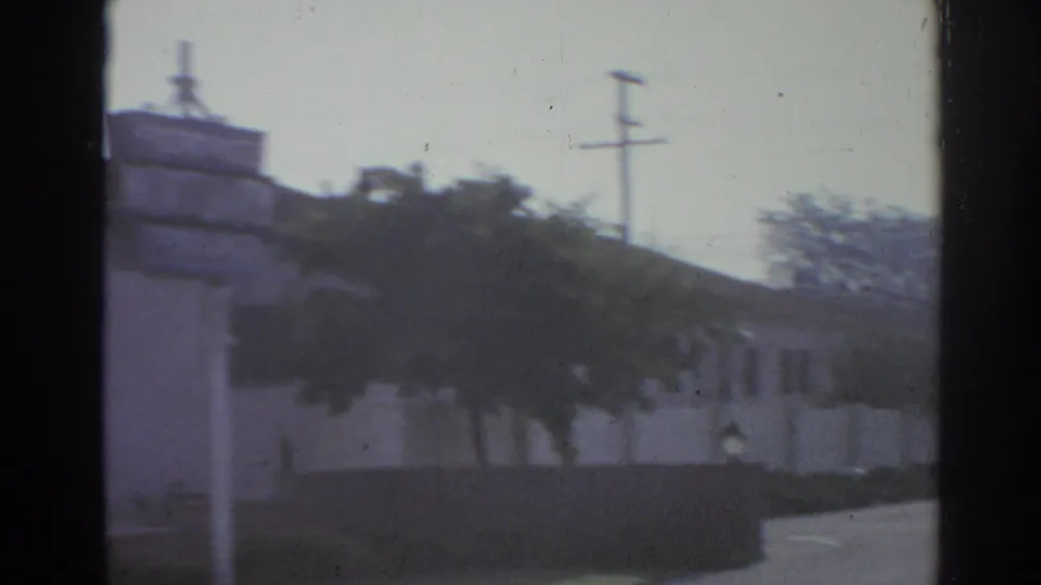 1966 San Gabriel street frame with a low commercial building, trees, utility pole, and wall seen from inside a moving vehicle.