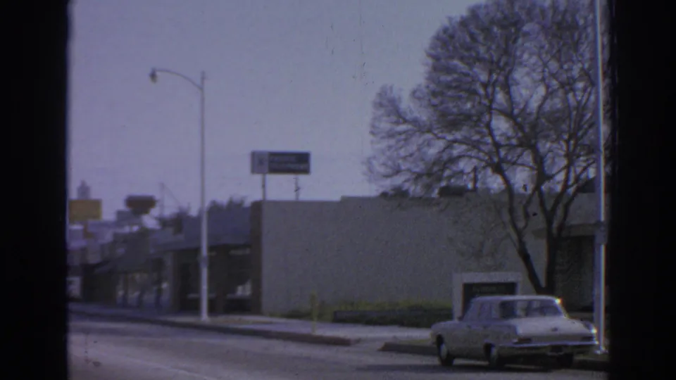 1966 San Gabriel drive-by frame showing a white sedan near low storefronts, streetlights, road signs, and a bare tree.