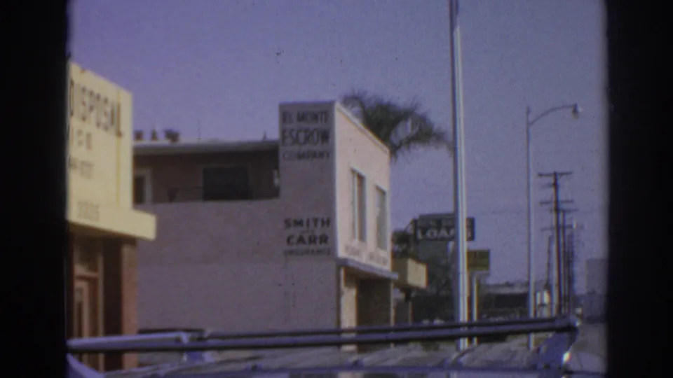 1966 San Gabriel frame showing mortuary and El Monte Escrow Company signage on roadside commercial buildings.