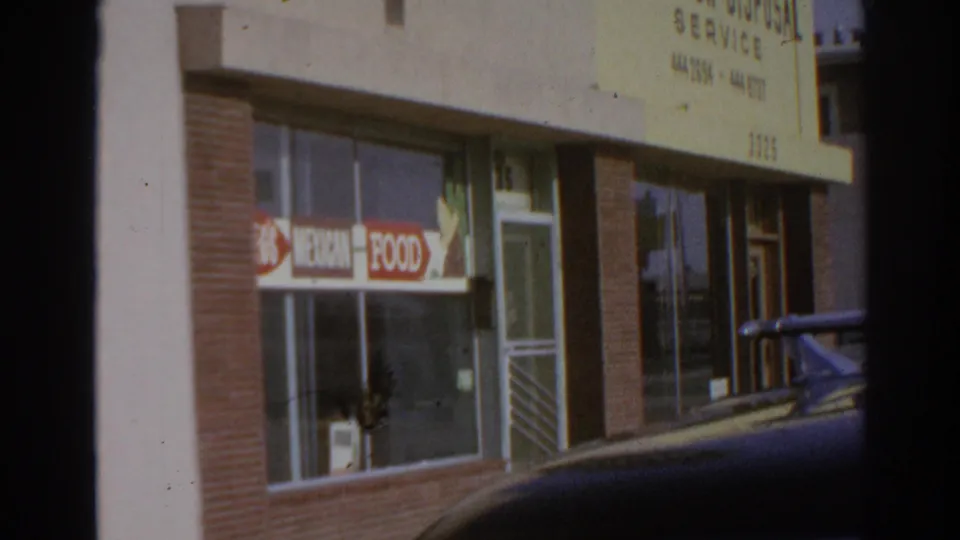 1966 San Gabriel storefront view with Anderton Disposal Service lettering, food signs in the windows, and a dark parked car along the curb.