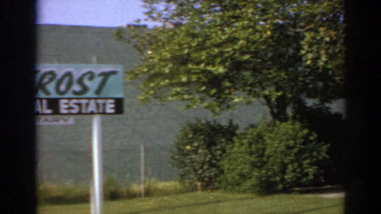 Real estate-notary sign beside trees and a pale building wall.