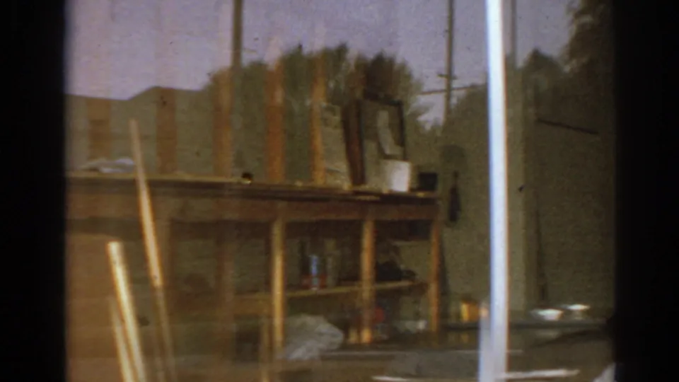 1966 San Gabriel garage or shed view with a wooden workbench, upright boards, shelves, containers, and a bright exterior edge.