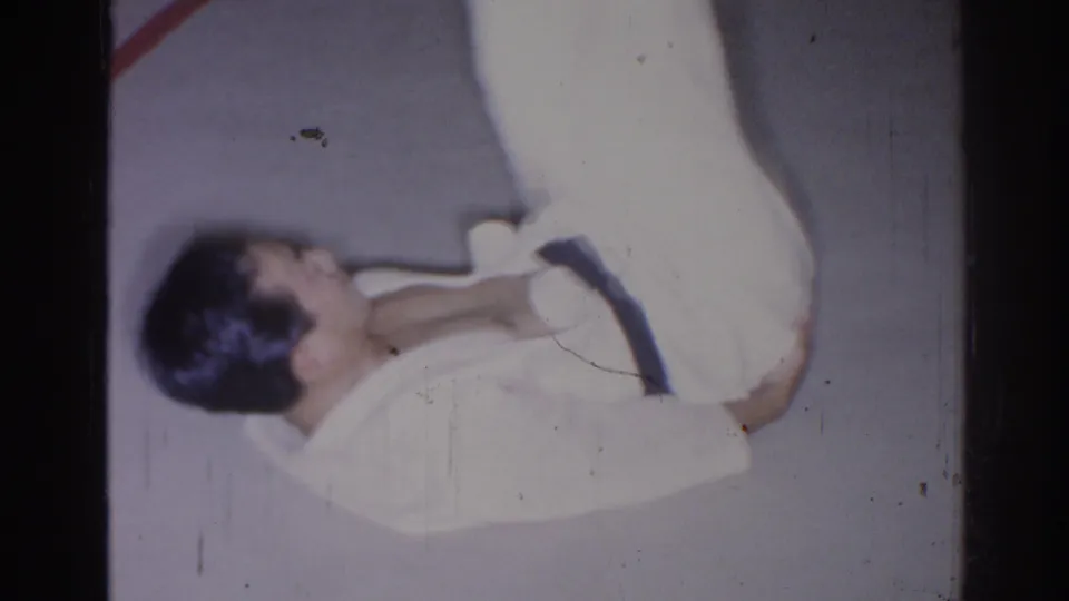 1966 San Gabriel frame showing an adult man in a white martial-arts gi lying on a pale indoor floor or mat.