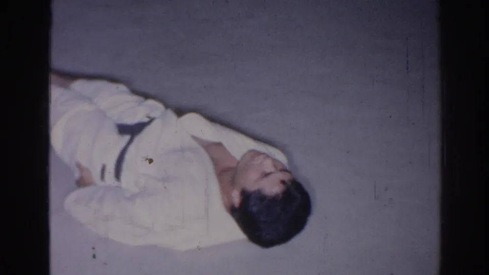In this 1966 San Gabriel 8mm frame, an adult man in a white gi lies sideways on concrete during floor exercise practice.