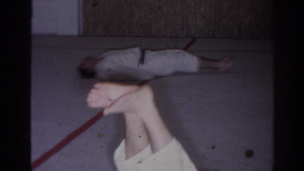 In this 1966 San Gabriel 8mm frame, the man in a white gi performs a leg raise on concrete beside a red painted line.