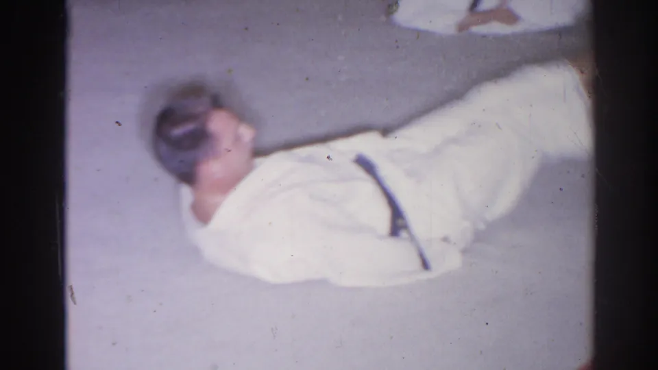 In this 1966 San Gabriel 8mm frame, the man continues a floor exercise, lifting one leg while lying on the concrete surface.