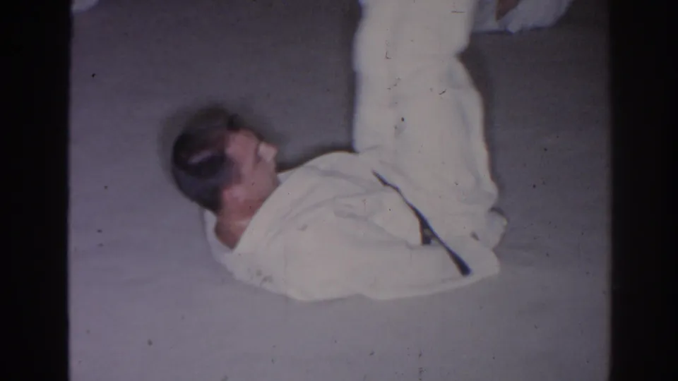 In this 1966 San Gabriel 8mm frame, an adult man in a white gi raises both legs while lying on concrete during floor training.