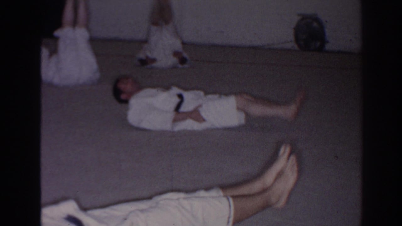 Students in white gi lie on a dojo mat while another person stands near the back wall.