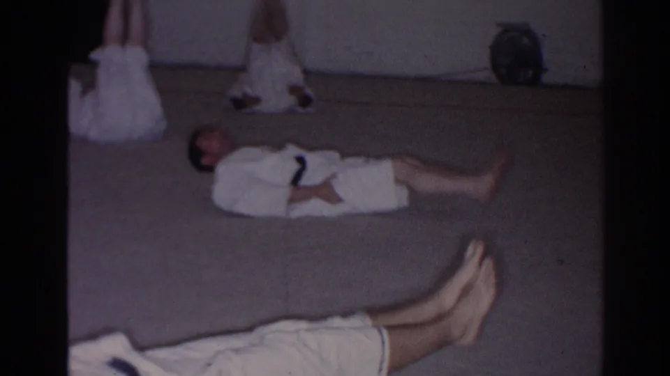 San Gabriel 1966 home-movie frame showing martial arts students in white gi resting or practicing floor positions on mats.