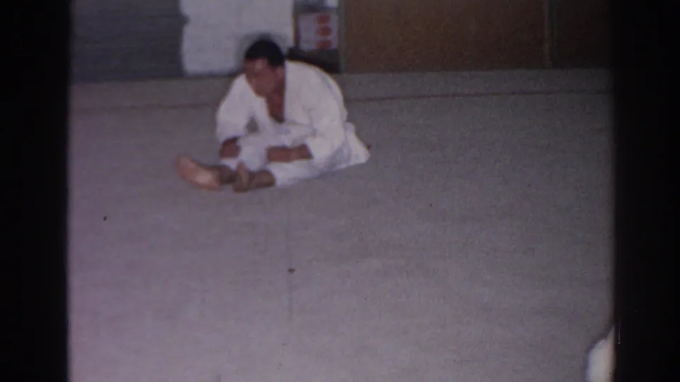 In 1966 San Gabriel footage, a martial arts practitioner in a white gi sits forward on a mat in a simple indoor room.