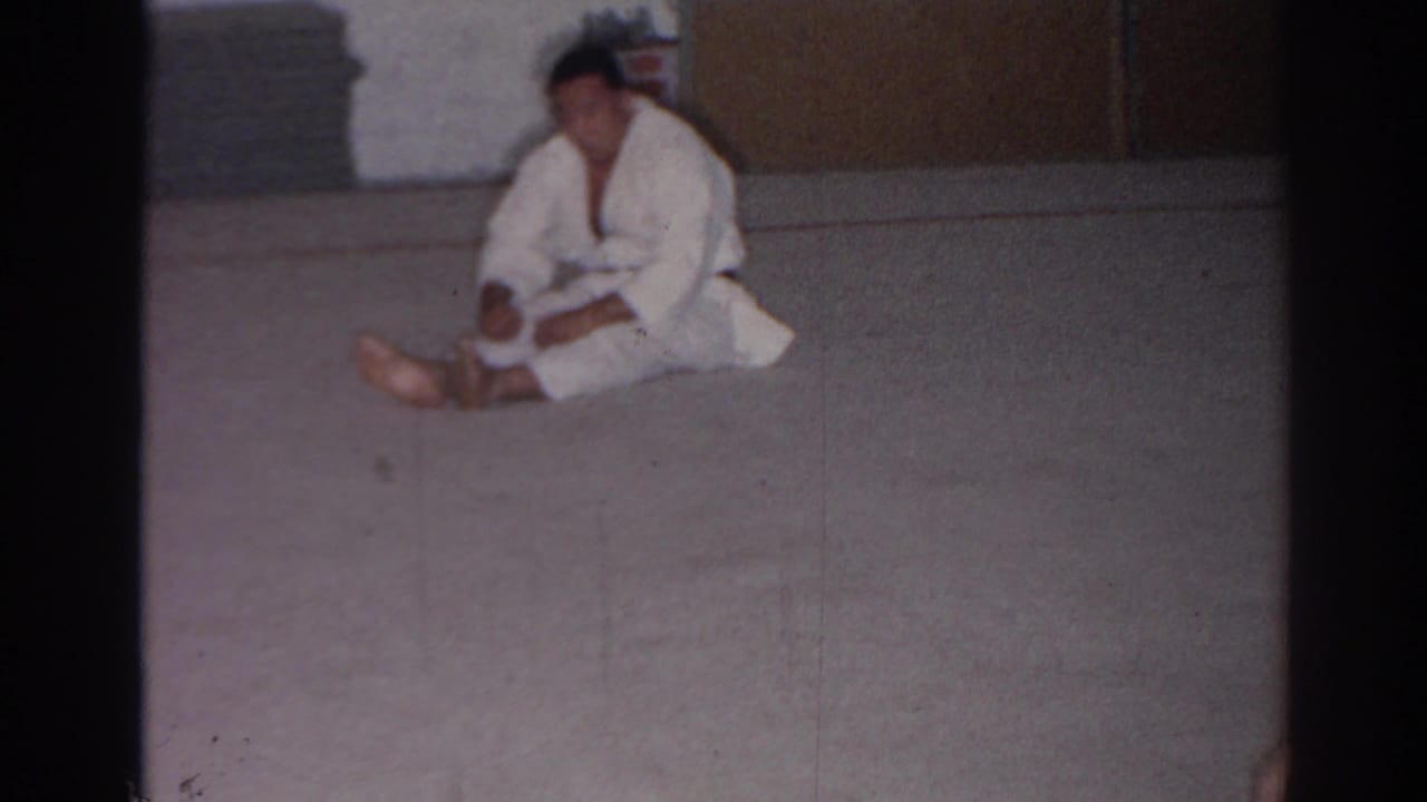 Adult man in a white karate-style uniform sits on the carpet inside a plain San Gabriel training room in 1966 footage.