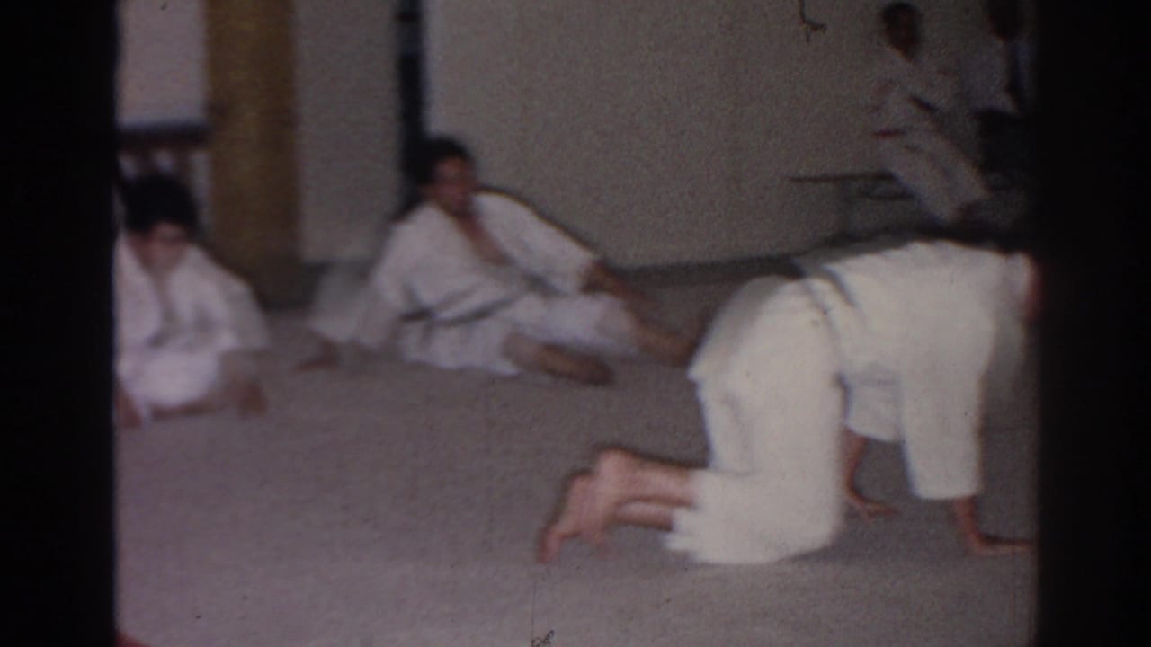 Men in white karate-style uniforms sit, kneel, and stretch on a carpeted floor during a 1966 San Gabriel training session.